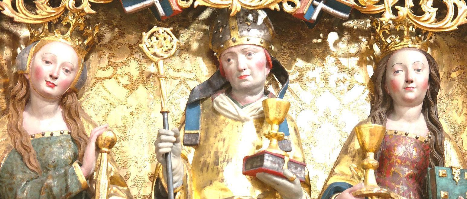 Two sculptures of humans with light skin tones, crowns, and long gold robes in front of an ornate gold background; figure on the left has short red hair and is holding a staff, gold cup, and book, and the figure on the right has long brown hair and is holding a gold cup and book