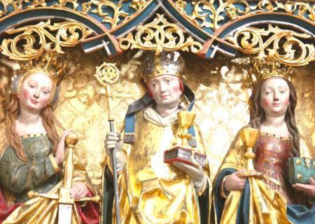 Two sculptures of humans with light skin tones, crowns, and long gold robes in front of an ornate gold background; figure on the left has short red hair and is holding a staff, gold cup, and book, and the figure on the right has long brown hair and is holding a gold cup and book