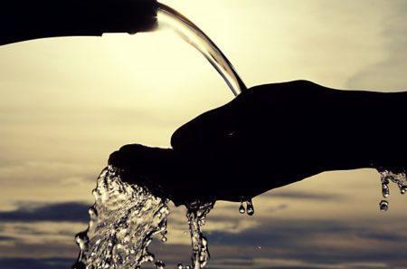 Silhouette of hand cupping water as it pours from a faucet in front of a blurred beige background