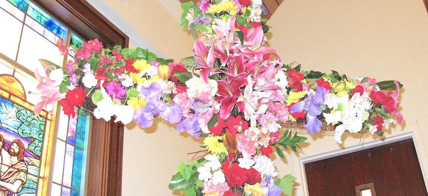 Cross covered in a variety of yellow, purple, red, white, and orange flowers and greenery in the corner of a room with a stained glass window