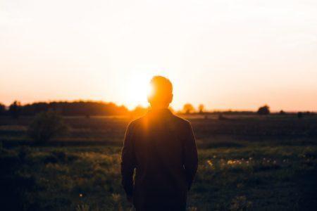 Back view of a person with short hair and long-sleeved shirt standing in a field in front of a shining sun