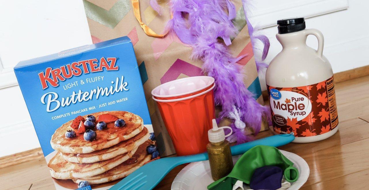 Box of pancake mix, red plastic cups, a blue plastic spatula, a bottle of maple syrup, a gift bag with a purple feather boa, and a white paper plate with uninflated balloons in various colors