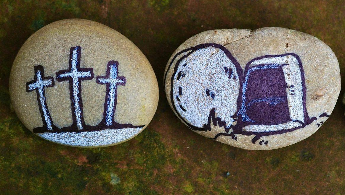 Close-up of two light brown rocks side by side on a green and brown background; the rock on the left is painted with three crosses in indigo and white, and the rock on the right is painted with an empty tomb and stone in indigo and white