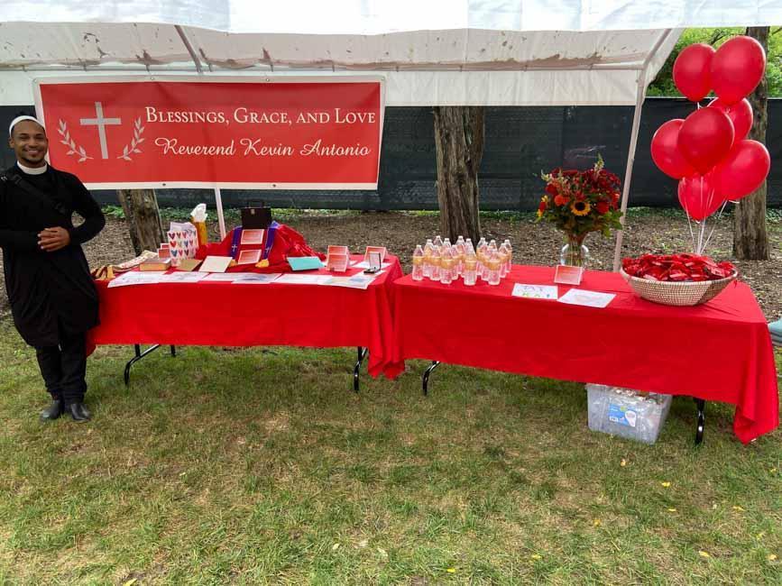 Priest standing by table with red cloth and balloons and give aways.
