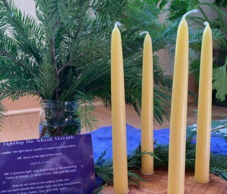 Wooden Advent wreath with beeswax candles, greenery in background, and prayer for lighting candles.