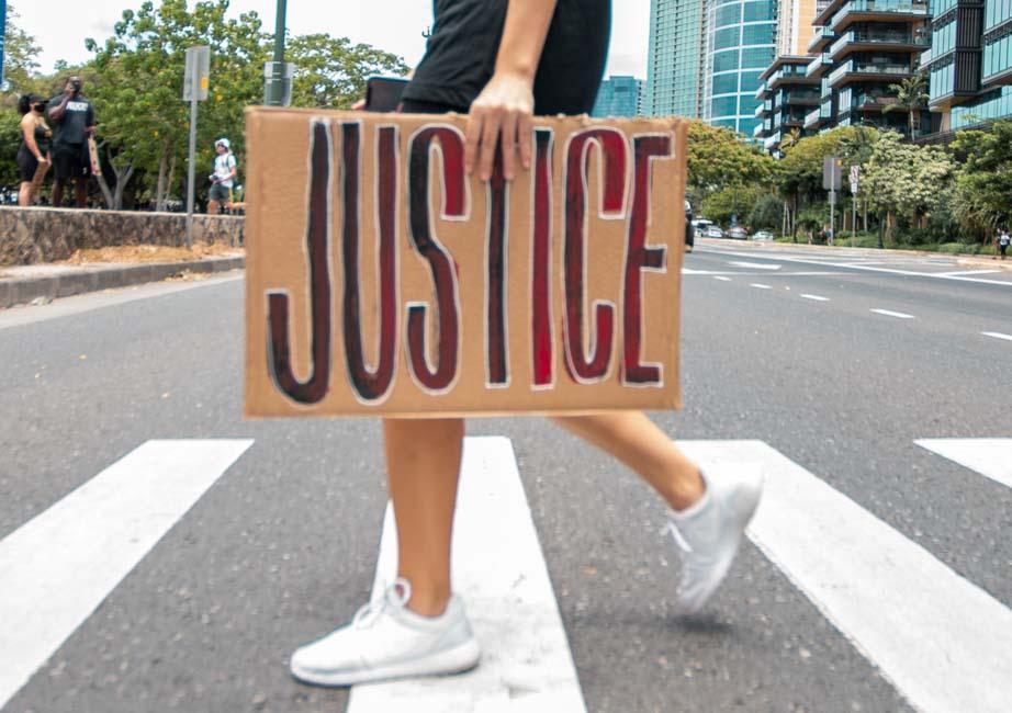 Cardboard sign with word "justice" painted in red and black. Sign in being carried across a cross walk by a white person.