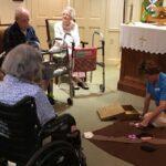 Older adults in chairs and wheelchairs watching woman on floor with Godly Play parable.