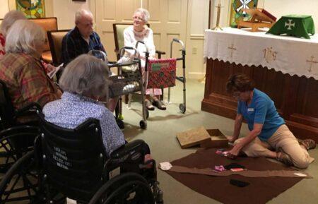 Older adults in chairs and wheelchairs watching woman on floor with Godly Play parable.