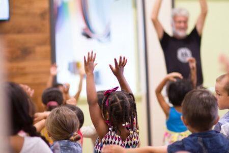 A group of children from behind with hands in the air and facing an older white man with raised hands.