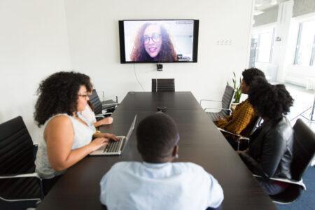 A small group of people at a table with computers and another person on a large screen.