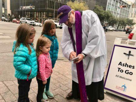 Three young children and a priest in front of an "Ashes To Go" sign.