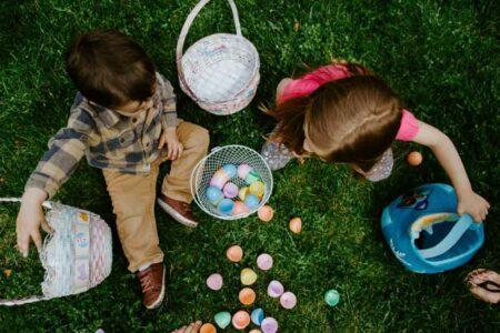 Aerial view of two children with brown hair and light skin tones sitting on grass with baskets of plastic Easter eggs