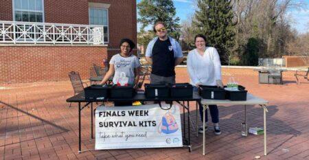 Three adults standing behind a table with a sign that reads "finals week survival kits."