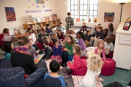 Woman sitting in chair with book surrounded by children.