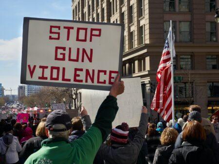 Crowd of individuals holding signs including one reading "Stop Gun Violence" and someone carrying an American flag.