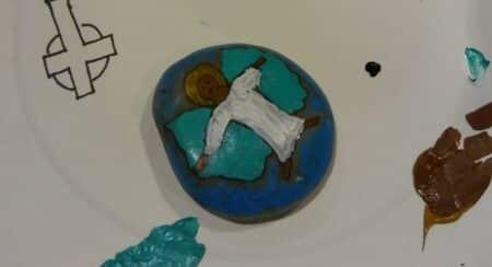 Rock painted with human figure with dark skin tone, white robe, and golden halo surrounded by paint samples and drawn crosses on a white paper plate