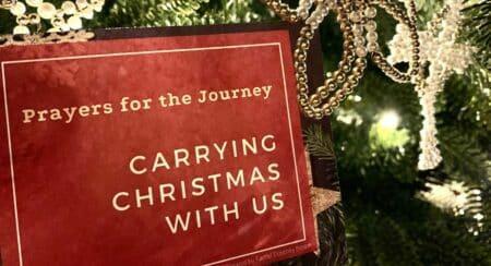 Close-up of red card with cream-colored text "Prayers for the Journey" and "Carrying Christmas with Us" in front of a decorated Christmas tree
