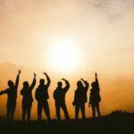 An orange sunset scene with six people in black silhouette standing in the foreground, each with a hand raised over their head