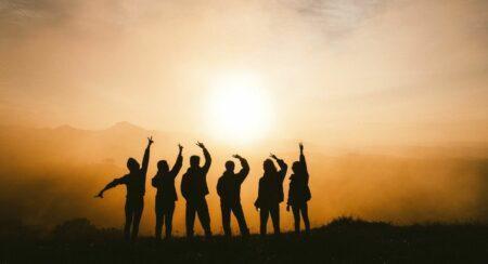 An orange sunset scene with six people in black silhouette standing in the foreground, each with a hand raised over their head