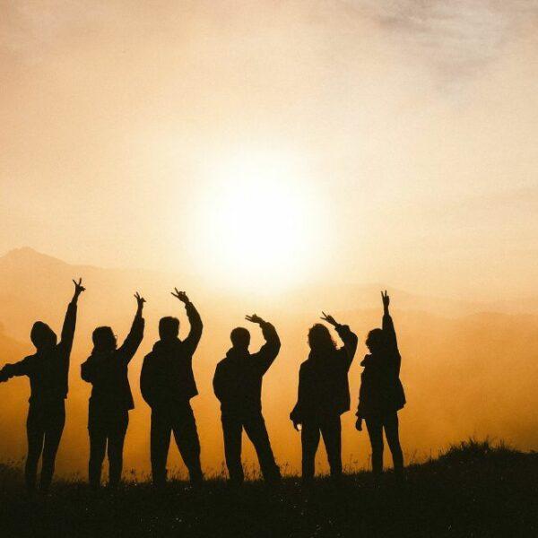 Silhouettes of six people standing with hands raised above their heads and sun in background