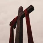 A view from below of a dark brown wooden cross with a red cloth draped over the horizontal beams against a cloudy background