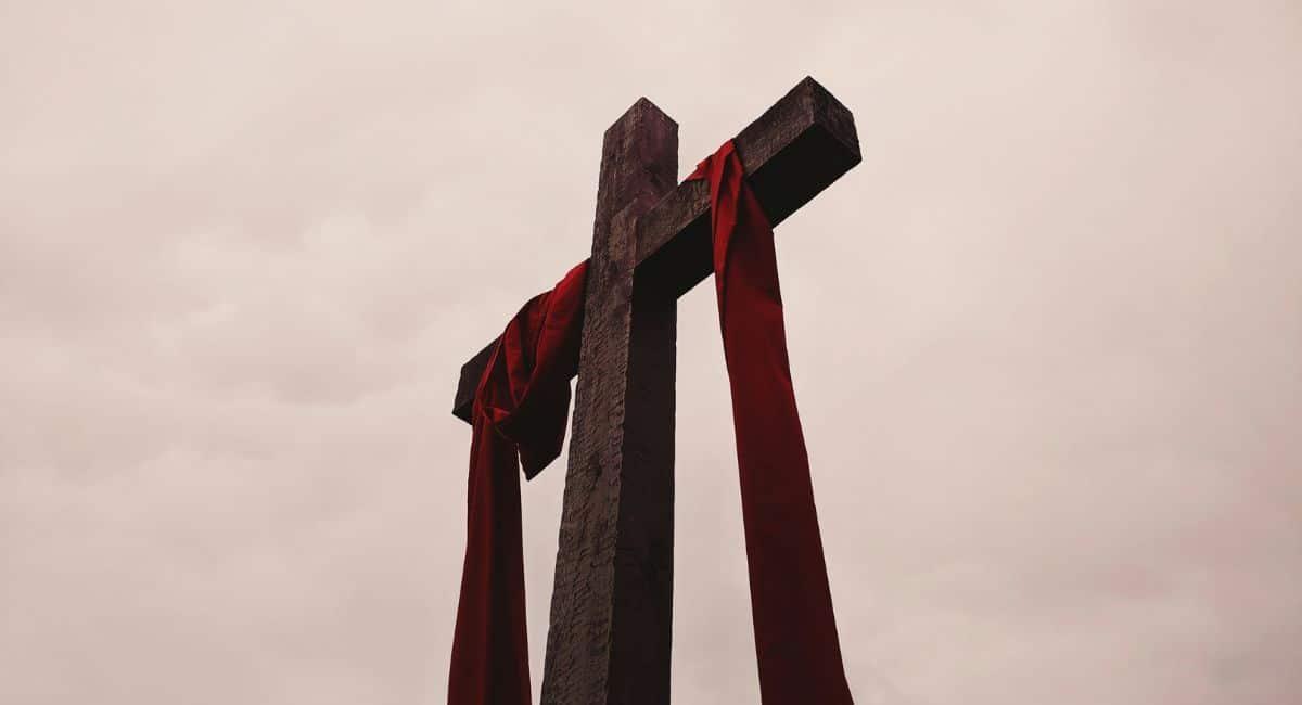 A view from below of a dark brown wooden cross with a red cloth draped over the horizontal beams against a cloudy background