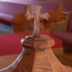 Close-up of a baptismal font's wooden cover with a cross at the top and purple cushioned pews in the background
