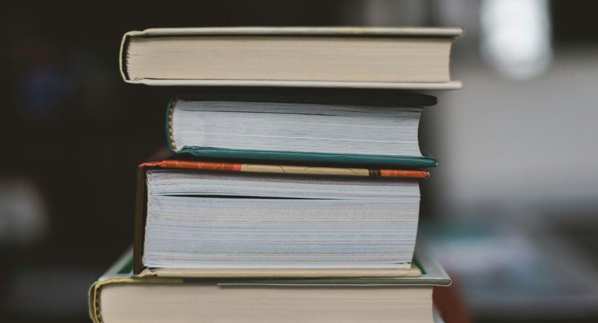 An end view of four thick hardback books stacked on top of one another with (in order from top to bottom) ivory, teal, red and beige, and yellow covers in front of a blurred background in dark and light gray