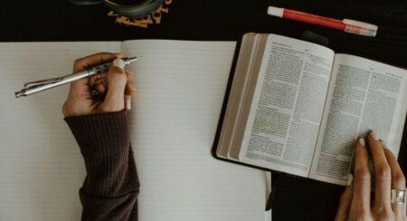 Aerial view of a person's hands, one holding a silver writing utensil over an open lined notebook and the other resting on an open Bible, with a mug and red marker nearby on top of a dark background; the person has brown skin and is wearing a dark brown long-sleeved top