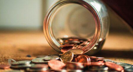 Close-up of a glass jar with a view from its mouth lying sideways on a tan-colored floor with numerous coins inside the jar and in front of it