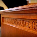 Angled view of a brown wooden table carved with the words "THIS DO IN REMEMBRANCE OF ME" with a black grand piano, gray floor, and beige wall in the background