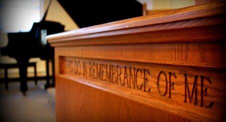 Angled view of a brown wooden table carved with the words "THIS DO IN REMEMBRANCE OF ME" with a black grand piano, gray floor, and beige wall in the background
