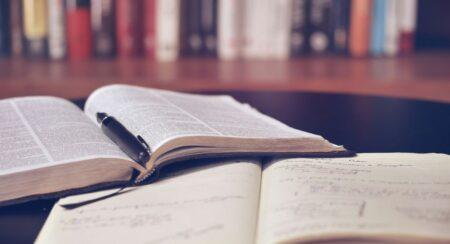 An open Bible with a black pen between the pages on top of an open notebook resting on a dark brown surface with a row of books blurred in the background