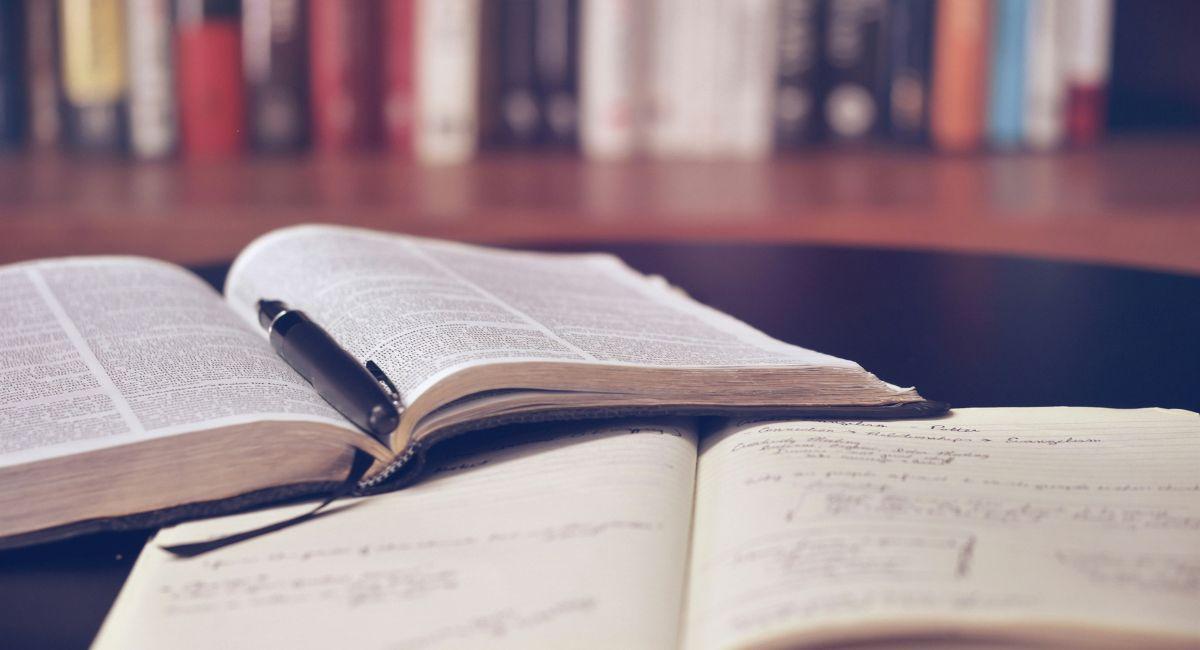 An open Bible with a black pen between the pages on top of an open notebook resting on a dark brown surface with a row of books blurred in the background
