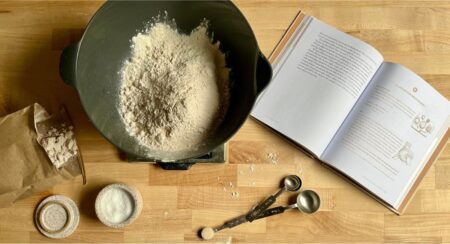 Aerial view of baking equipment and ingredients with a book open to "A Liturgy for Bread Baking" on a light brown wooden surface; items include (left to right, clockwise) a brown bag of flour, a dark bowl holding light brown flour, open book, 4 metal measuring spoons with the smallest holding a light brown dry ingredient, and a ceramic jar filled with a white dry ingredient and ceramic lid