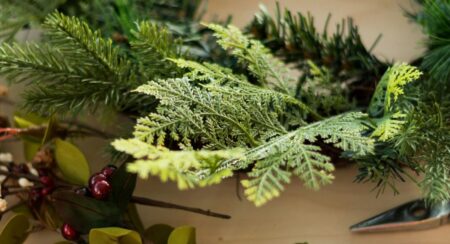 Close-up of evergreens gathered into a wreath with a bundle of red berries and the tip of a pair of metal pliers on a beige surface