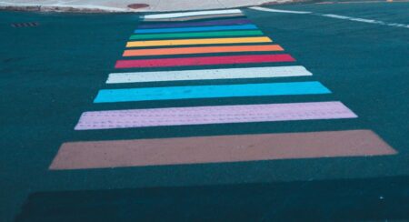 Street painted with stripes in colors of the Pride flag, from top to bottom: white, brown, purple, blue, green, yellow, orange, red, white, light blue, light pink, brown, and black