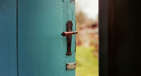 Close-up of blue-green door with copper-colored handle and metal latch opening to an outdoor landscape, which is blurred in the background
