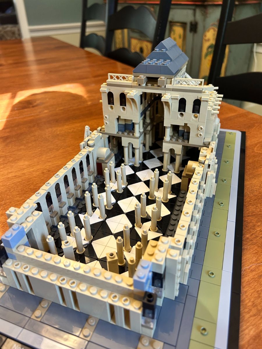 Angled aerial view of interior floor and pillars of Lego Notre Dame Cathedral on a tan wood surface with black chairs and a blue-gray piece of furniture in the background