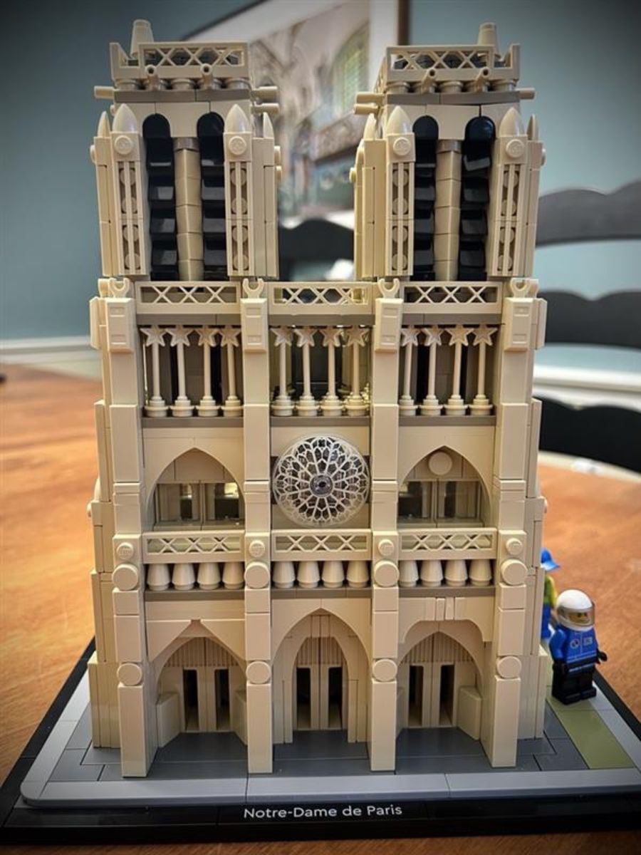 Side view of the two towers, rose window, and entrances of Lego Notre Dame Cathedral on a tan wood surface with black chairs, a teal wall, and a work of art in the background