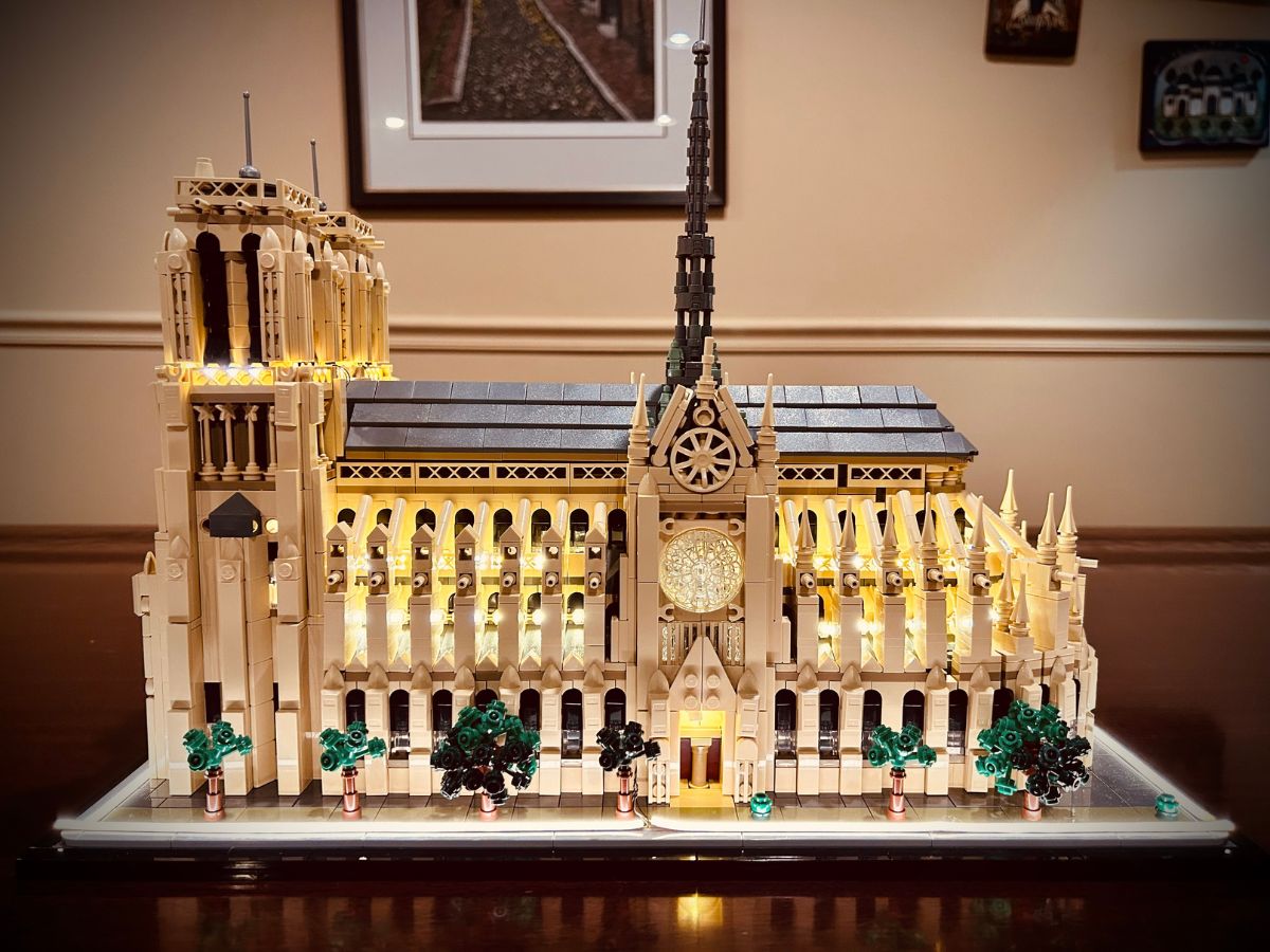 Side view of a fully constructed Lego Notre Dame Cathedral with electric lights on a flat surface with a beige and dark brown wall and partial view of three framed works of art in the background
