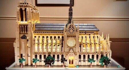 Side view of a fully constructed Lego Notre Dame Cathedral with electric lights on a flat surface with a beige and dark brown wall and partial view of a picture frame in the background
