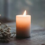 Lit pillar candle with peach and blue-gray hues on a flat gray surface with a branch to the left and windowpanes in the background