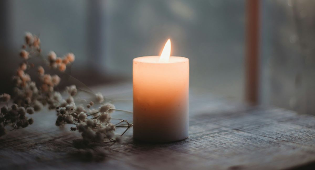 Lit pillar candle with peach and blue-gray hues on a flat gray surface with a branch to the left and windowpanes in the background