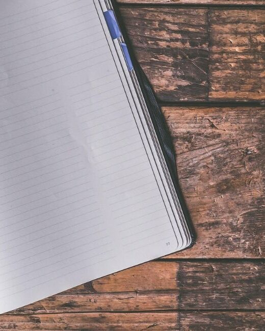 Partial view of an open lined notebook with white paper and a silver pen with a blue cap in the notebook crease on top of a dark brown wooden surface