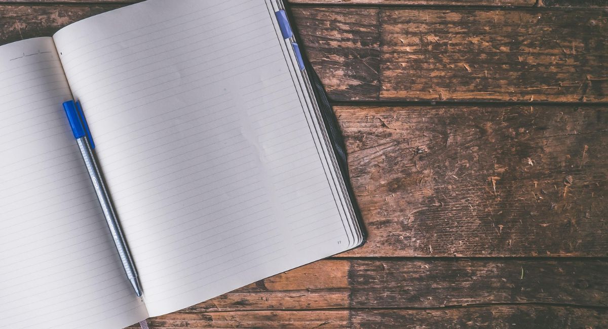Partial view of an open lined notebook with white paper and a silver pen with a blue cap in the notebook crease on top of a dark brown wooden surface