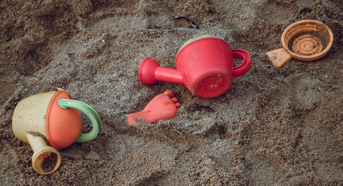 Plastic toys on wet brown sand; from left to right, a yellow, orange, and green water pitcher, an orange footprint mold, a red water pitcher, and an orange sieve