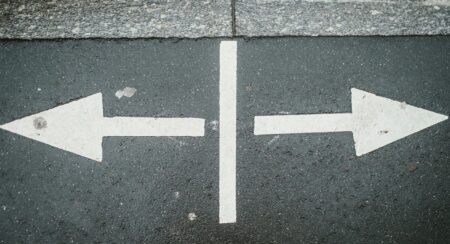Two white arrows pointing outward in opposite directions with a vertical white line between them on dark gray pavement