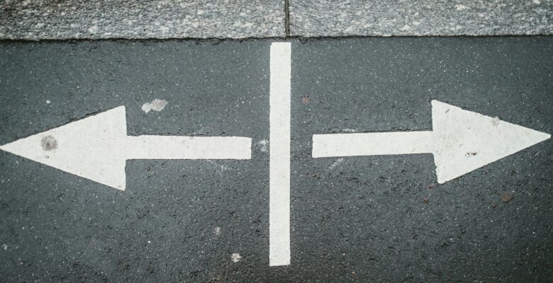 Two white arrows pointing outward in opposite directions with a vertical white line between them on dark gray pavement