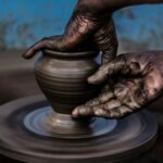 Two hands of a person with dark skin tone shaping bronze-colored clay on a pottery wheel in front of a blurred blue background with brown spots